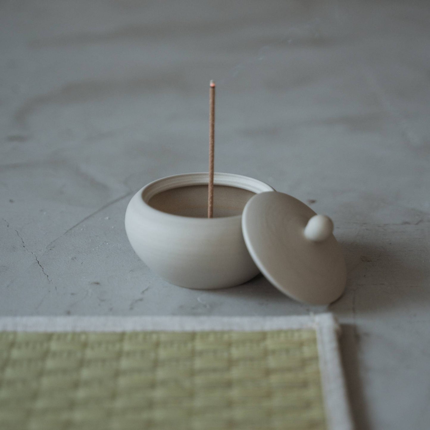 "Weißer japanischer Räucherstäbchenhalter in Form einer Mönchsschale mit brennendem Stäbchen und aufsteigendem Rauch in der Nähe einer Tatami-Matte."