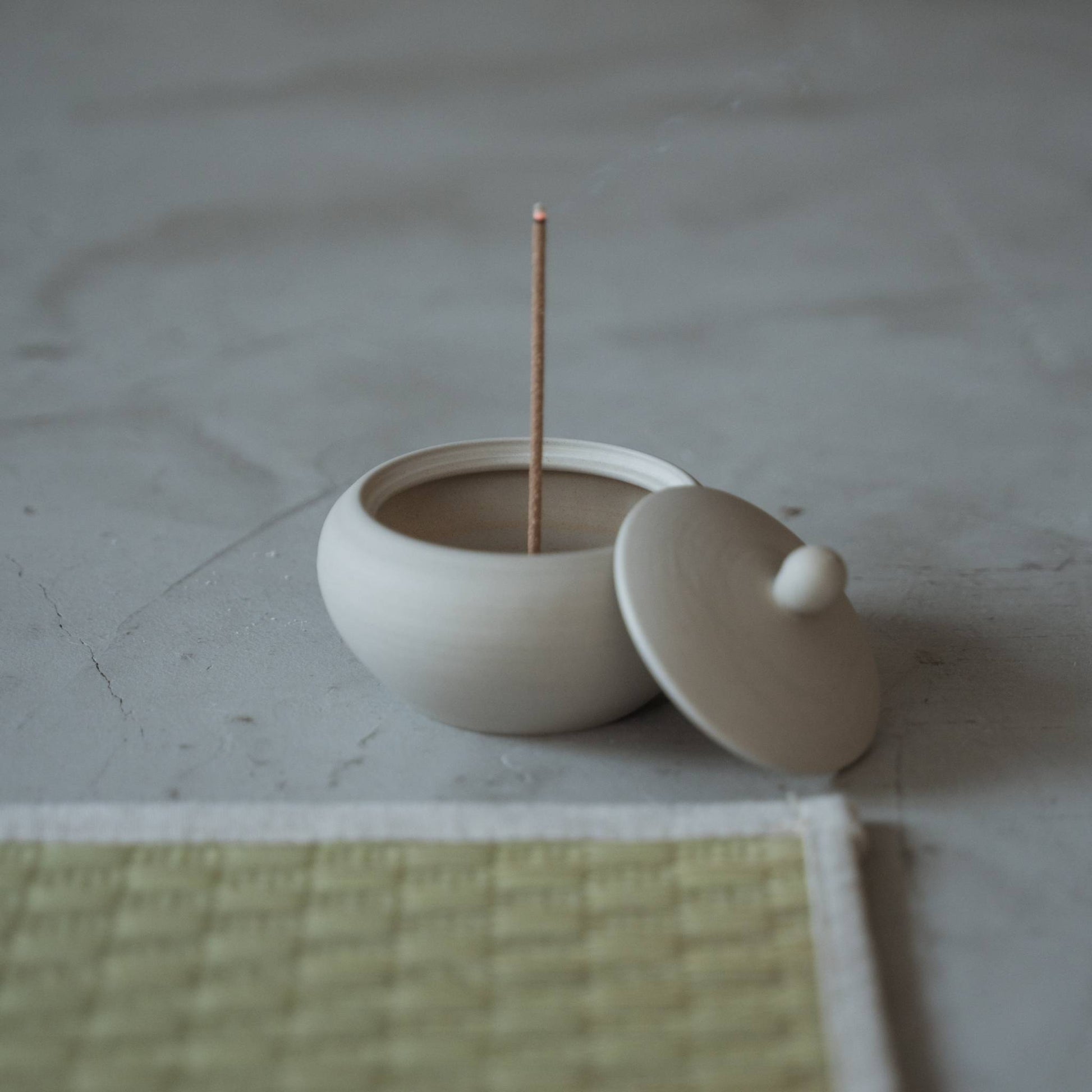 "Weißer japanischer Räucherstäbchenhalter in Form einer Mönchsschale mit brennendem Stäbchen und aufsteigendem Rauch in der Nähe einer Tatami-Matte."