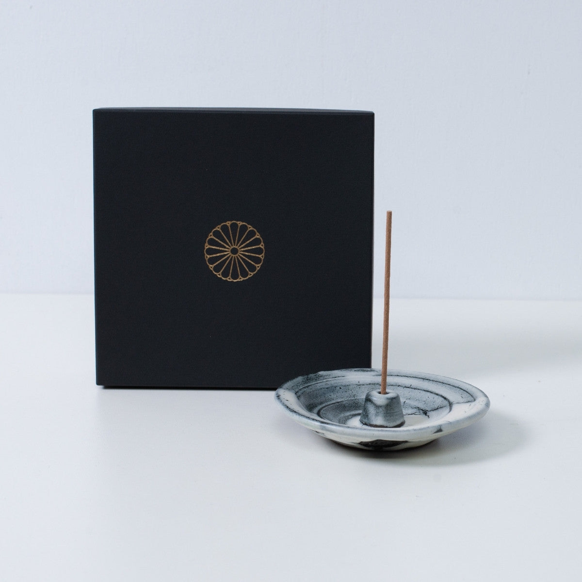 White and black Japanese incense holder "Creative Cloud" pottery with a stick next to a black gift box with a gold chrysanthemum crest.