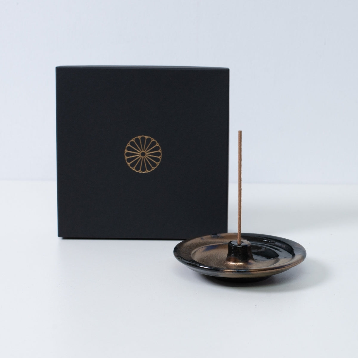 Goldfarbener und schwarzer japanischer Räucherstäbchenhalter "Zen" aus Keramik mit einem Stäbchen neben einer schwarzen Geschenkbox mit goldenem Chrysanthemenwappen, hergestellt in Japan.