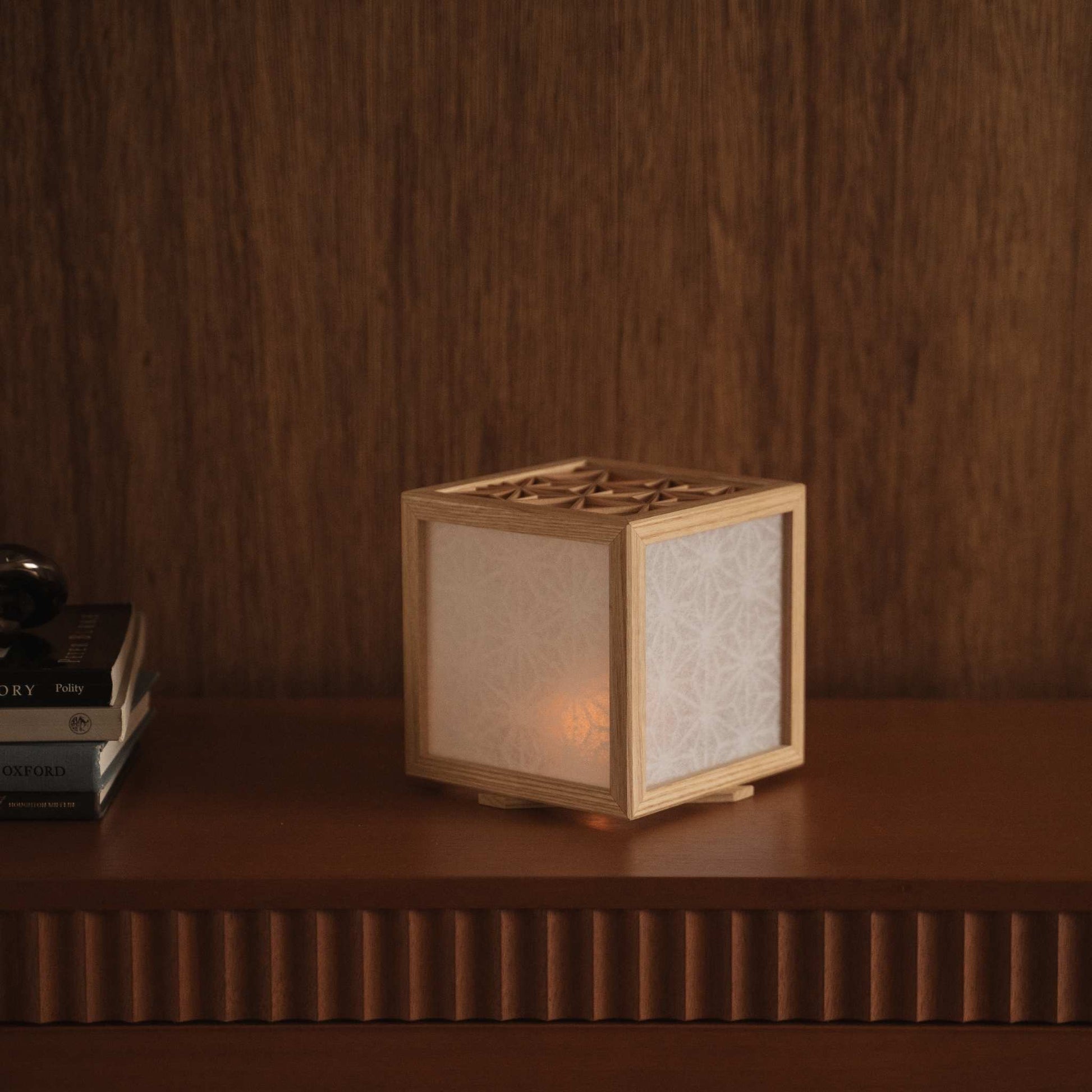 Glimrende japansk kumiko lampeskærm, der sidder på en mørk træhylde ved siden af bøger og skaber en varm og elegant atmosfære.