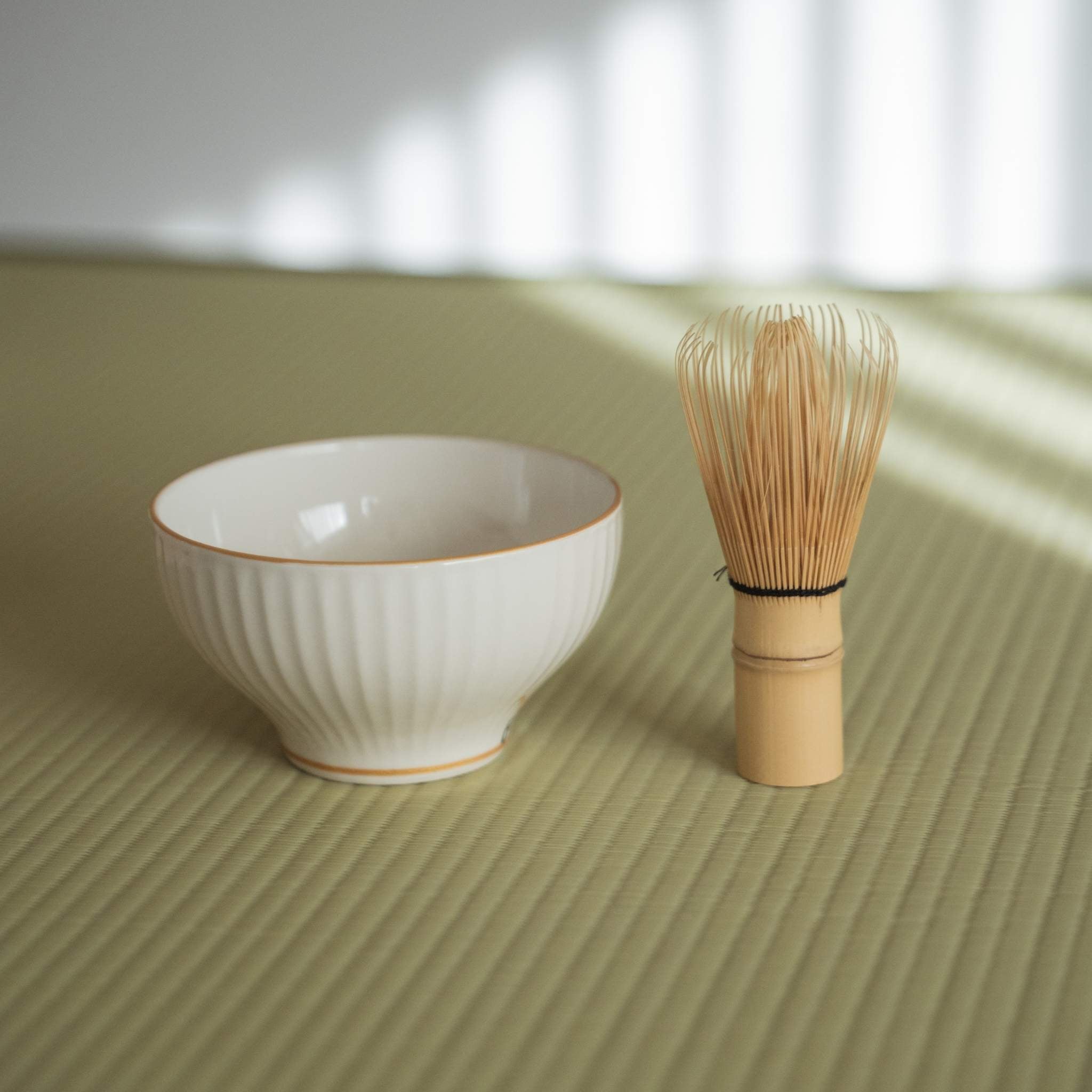 Sidevisning af en ribbet hvid porcelæns matcha-skål, der står på en traditionel tatami-måtte ved siden af et te-piskeris.