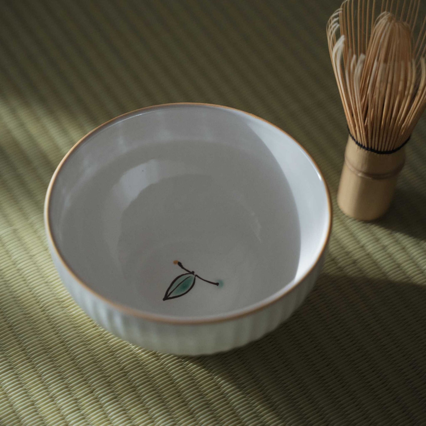 Indvendigt billede af en hvid japansk matcha-skål med en lille malet knop ved siden af en bambus-chasen på en tatami-måtte.