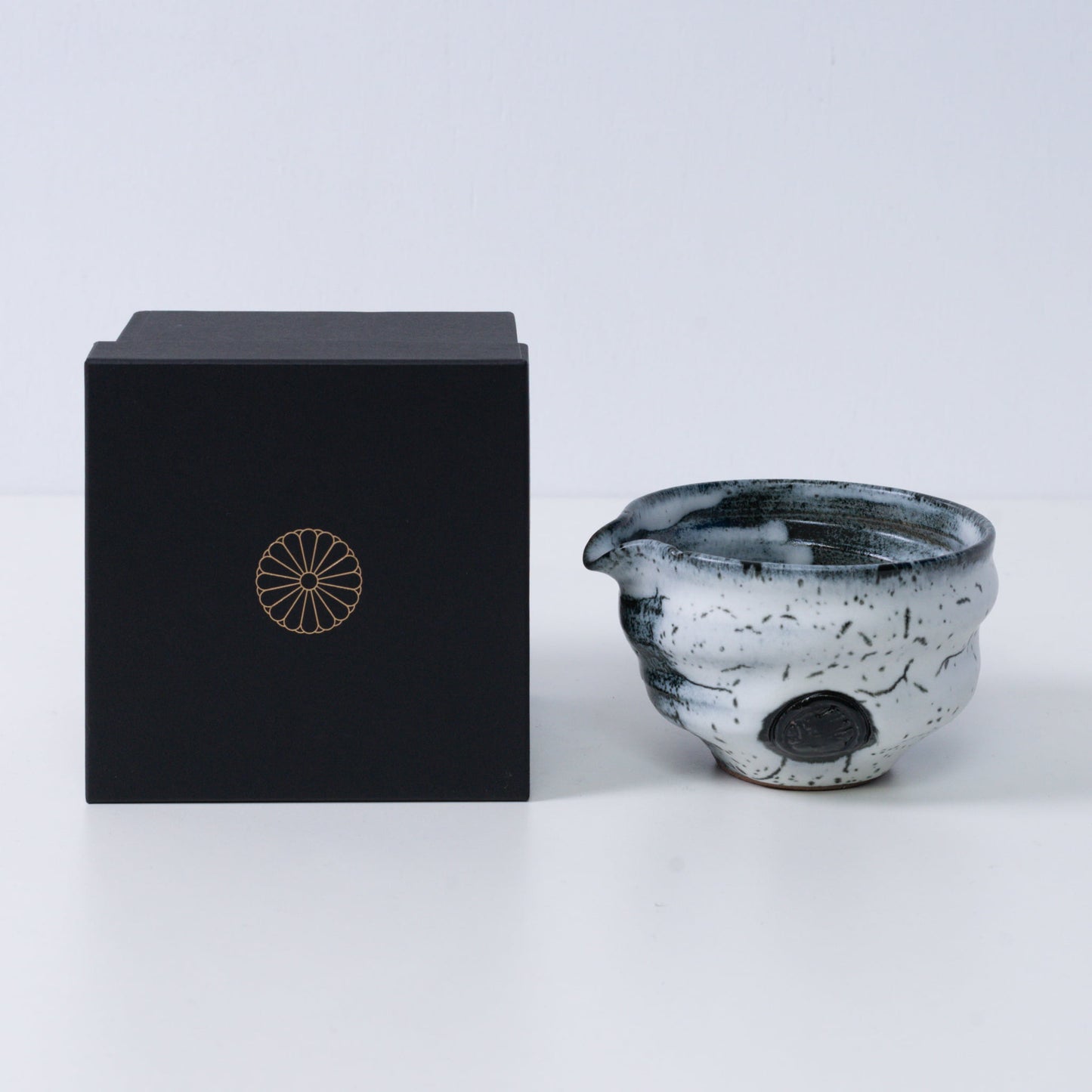 White and black Japanese matcha bowl with spout "Creative Cloud "pottery beside a black gift box with a gold chrysanthemum crest, from Japan.