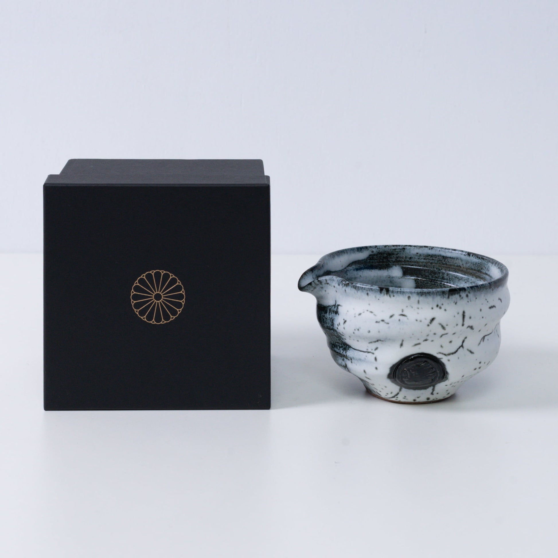 White and black Japanese matcha bowl with spout "Creative Cloud "pottery beside a black gift box with a gold chrysanthemum crest, from Japan.