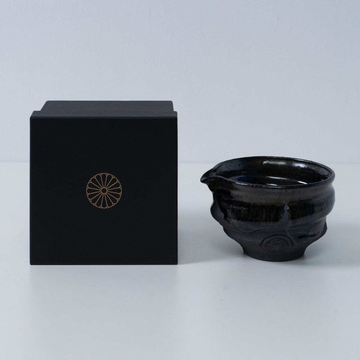 Japansk matcha-skål med tud "Dignity Blue" keramik ved siden af en sort gaveæske med et gyldent krysantemum-emblem, fra Japan.