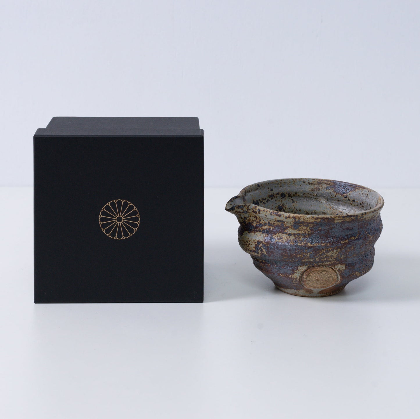 Rustic brown Japanese matcha bowl with spout "Sakigake" pottery next to a black gift box with a gold chrysanthemum crest, made in Japan.