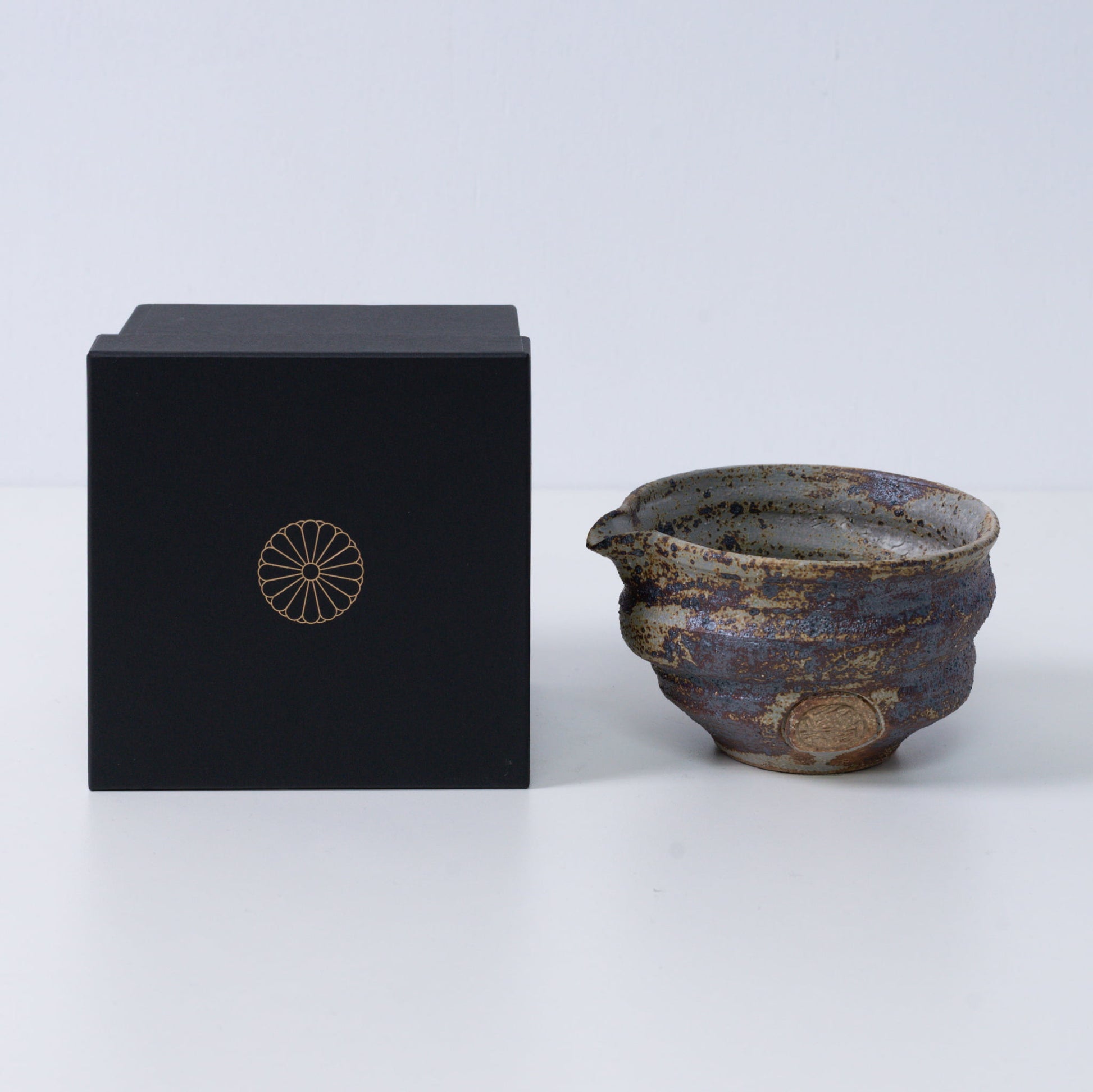 Rustic brown Japanese matcha bowl with spout "Sakigake" pottery next to a black gift box with a gold chrysanthemum crest, made in Japan.