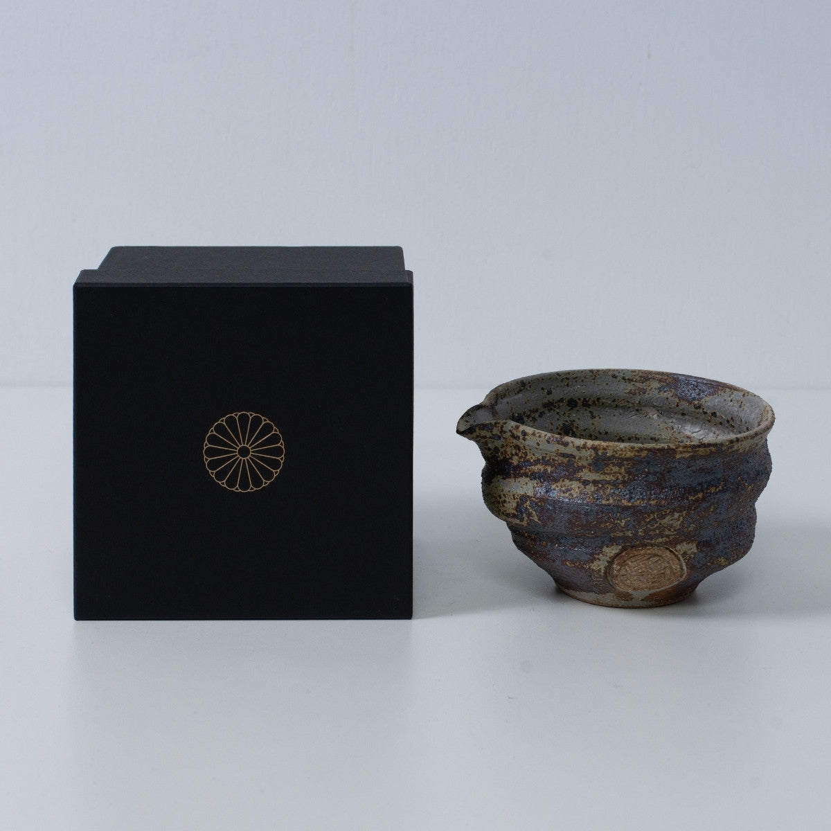 Rustik brun japansk matcha-skål med tud "Sakigake" keramik ved siden af en sort gaveæske med et gyldent krysantemum-emblem, lavet i Japan.