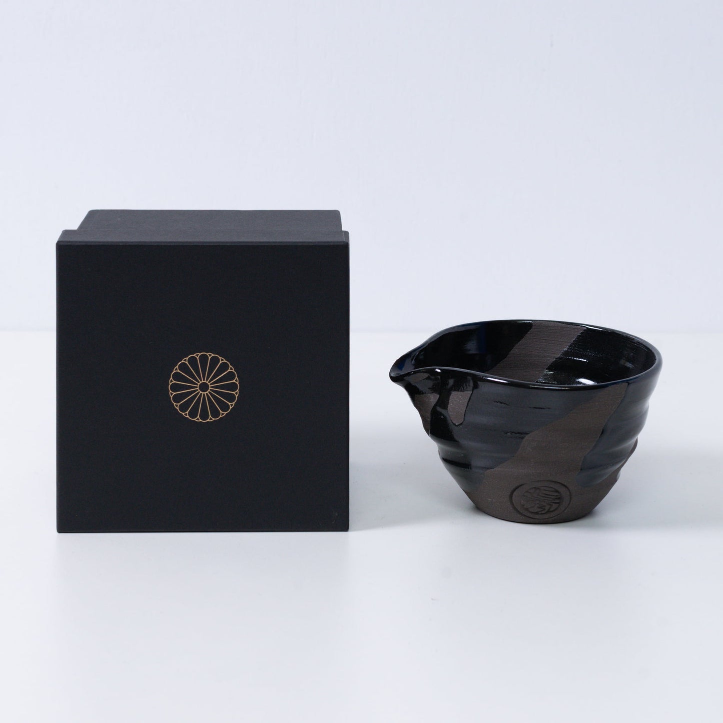 Japanese matcha bowl with spout "Sumi Black" pottery next to a black gift box with a gold imperial chrysanthemum crest, Kyoto ceramics.