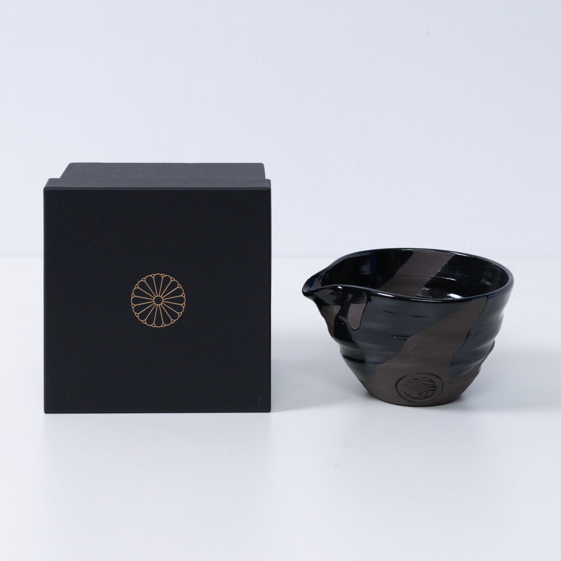 Japanese matcha bowl with spout "Sumi Black" pottery next to a black gift box with a gold imperial chrysanthemum crest, Kyoto ceramics.