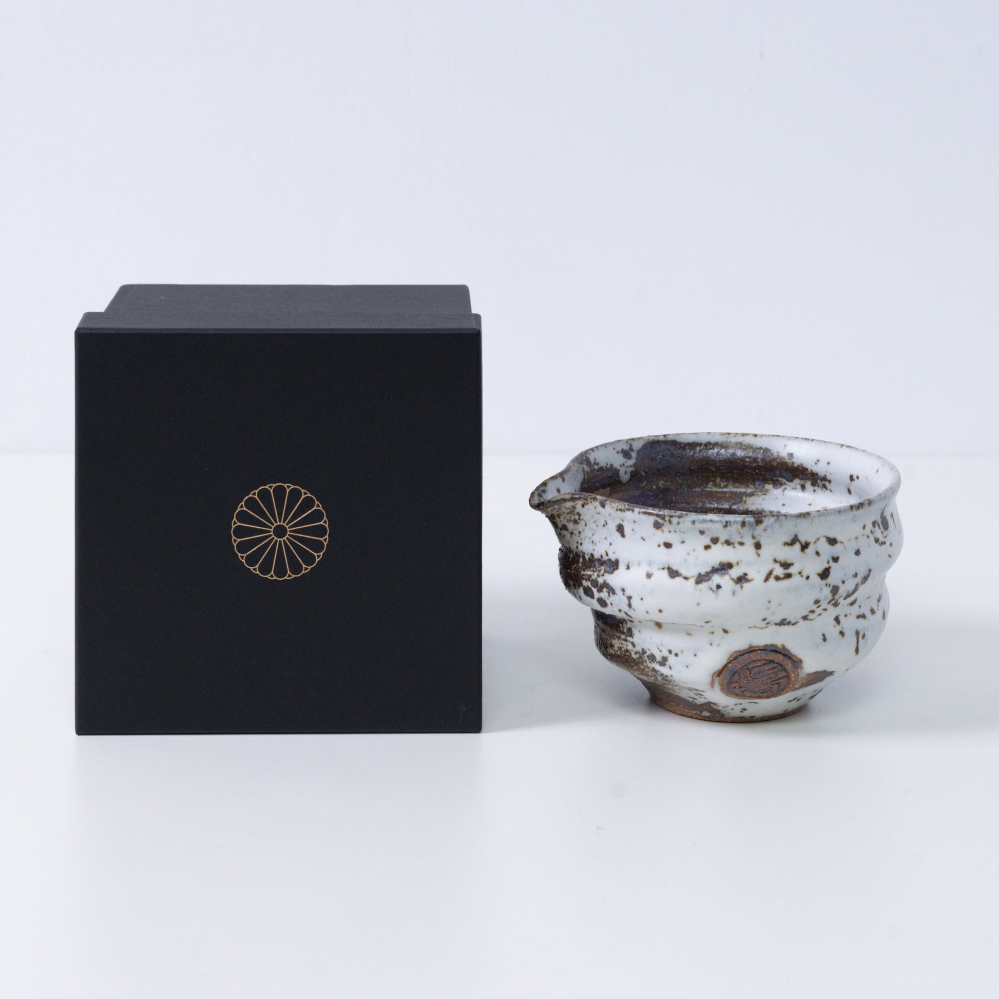 Brown and white Japanese matcha bowl with spout "Zui" pottery next to a black gift box with a gold imperial chrysanthemum crest, from Japan.