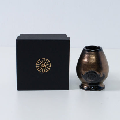 Support en céramique noir et or pour fouet à matcha japonais "Zen", à côté d'une boîte cadeau noire avec un blason de chrysanthème doré, du Japon.