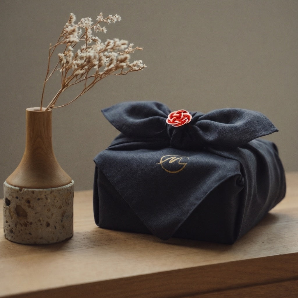 A front view photograph of a Japanese gift item wrapped in an authentic navy Furoshiki cloth on a wooden table, with the neat 'ma-musubi' knot centered.