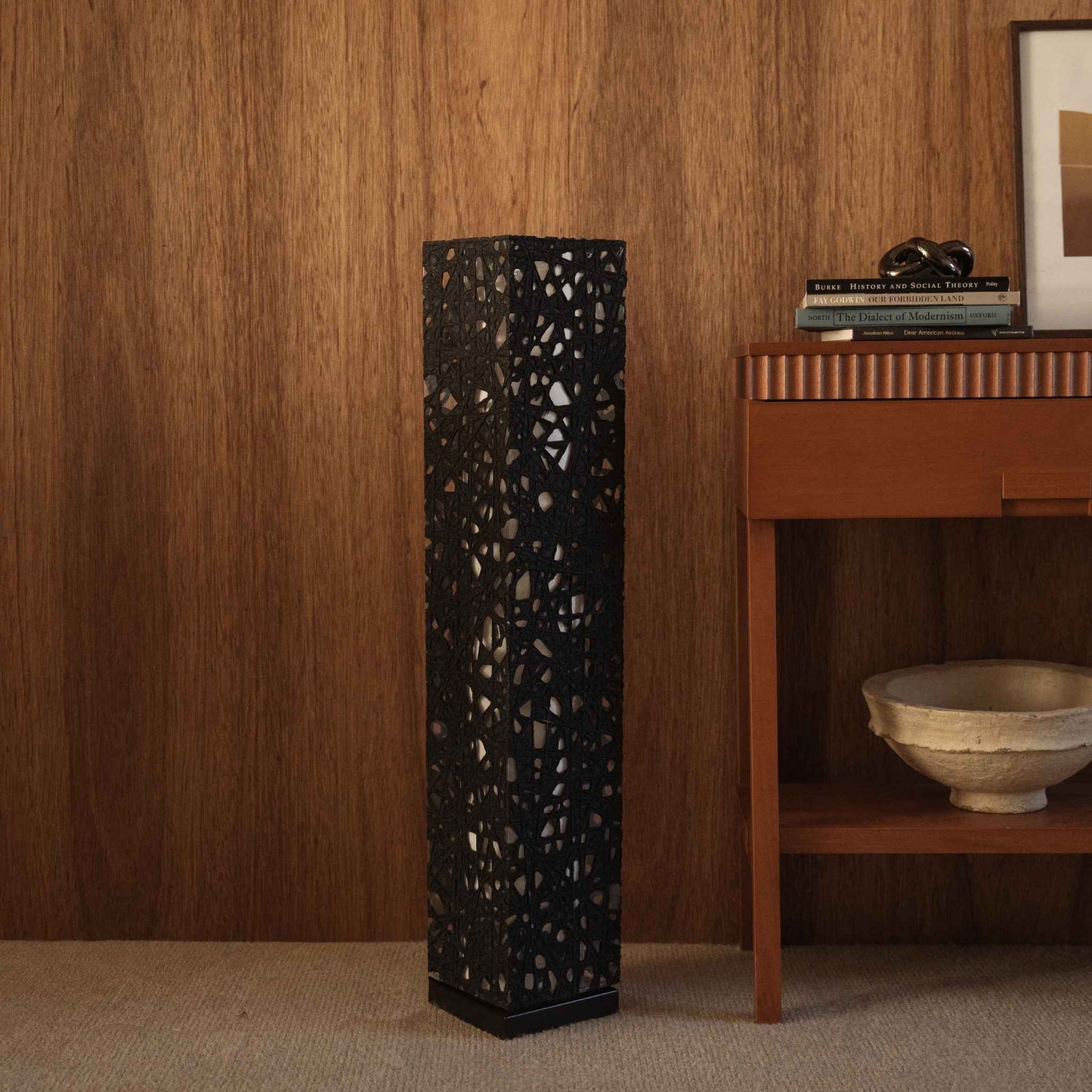 "Lampe sur pied noire Me Look 01 en papier, non allumée, posée sur un tapis devant un mur en bois sombre près d'une console."