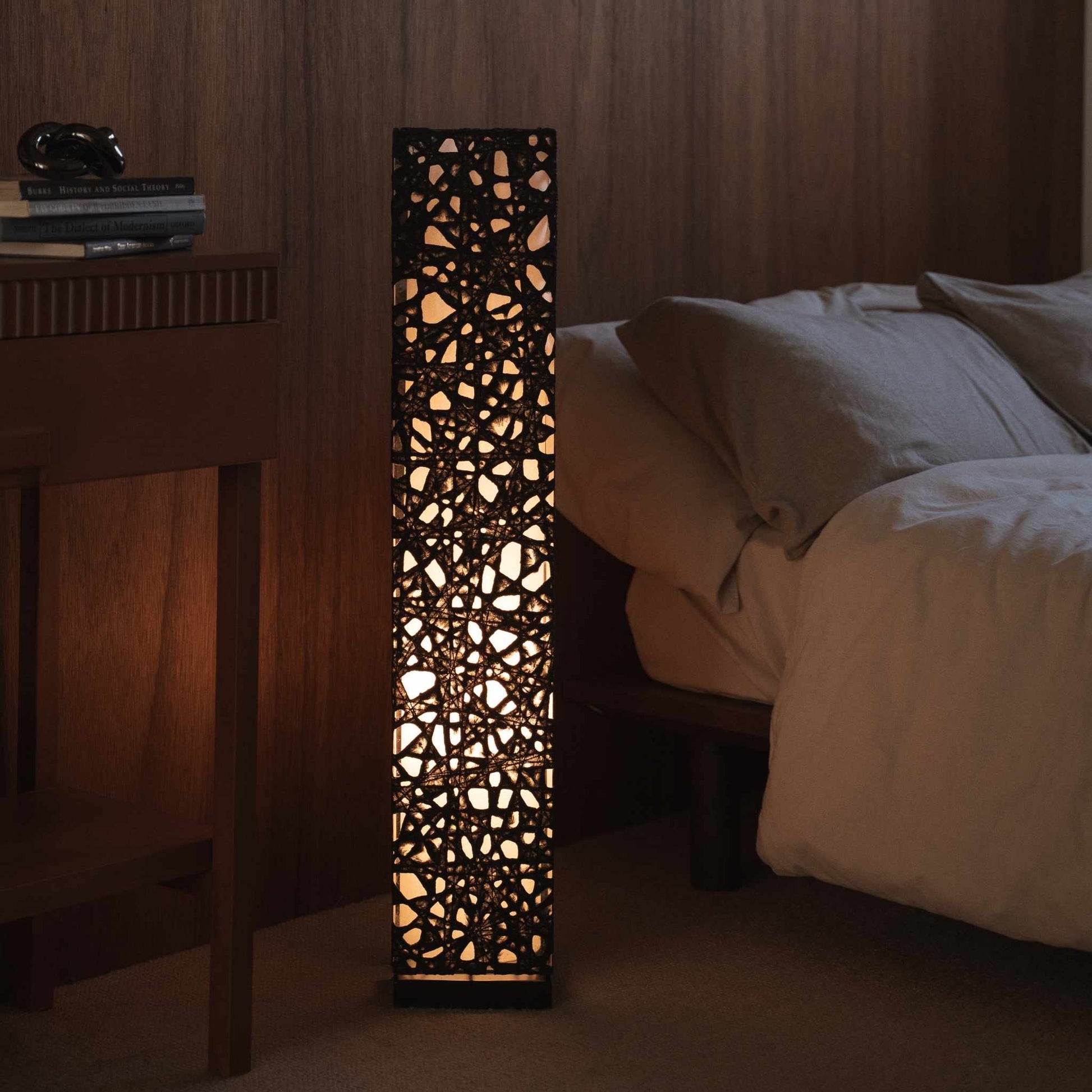 "Lampe sur pied en papier japonais noir Me Look 01, placée à côté d'un lit dans une chambre japonaise faiblement éclairée."