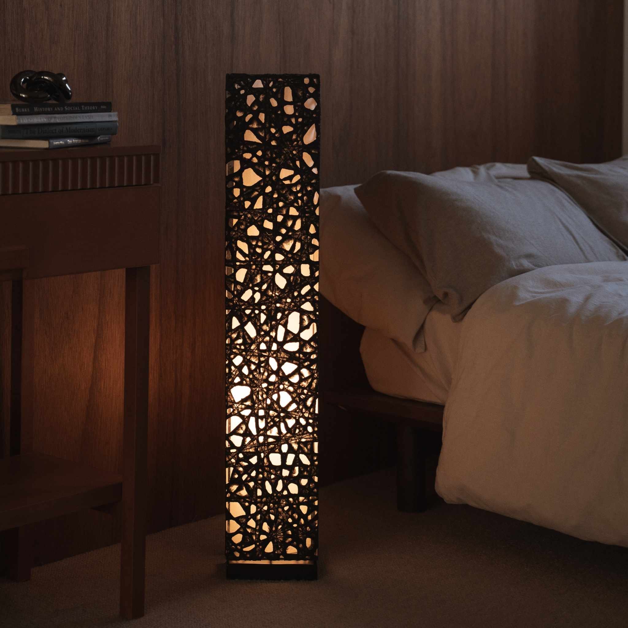 "Lampe sur pied en papier japonais noir Me Look 01, placée à côté d'un lit dans une chambre japonaise faiblement éclairée."