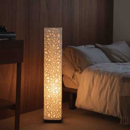 "Lampe sur pied japonaise Me Look 01 en papier blanc lumineux diffusant une lumière chaude à côté d'un lit dans une chambre japonaise."