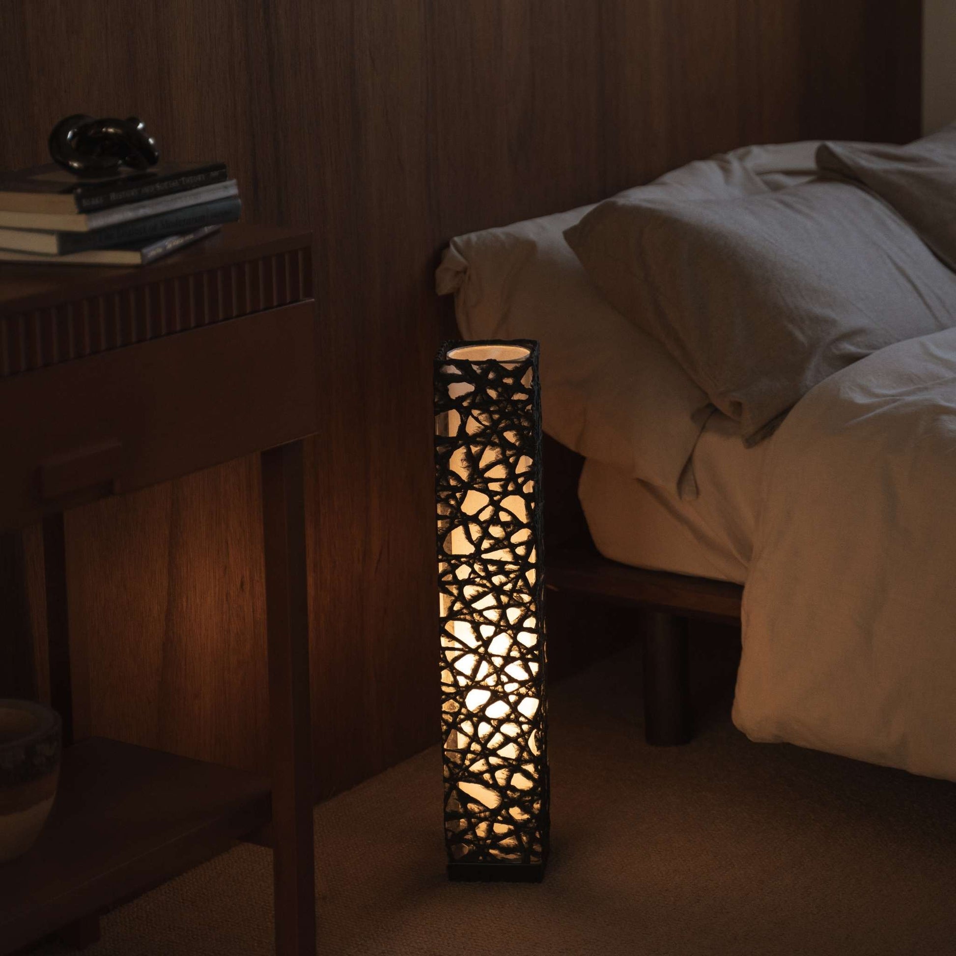 "Lampe sur pied en papier japonais noir Me Look 02, éclairant chaleureusement à côté d'un lit dans une chambre japonaise."