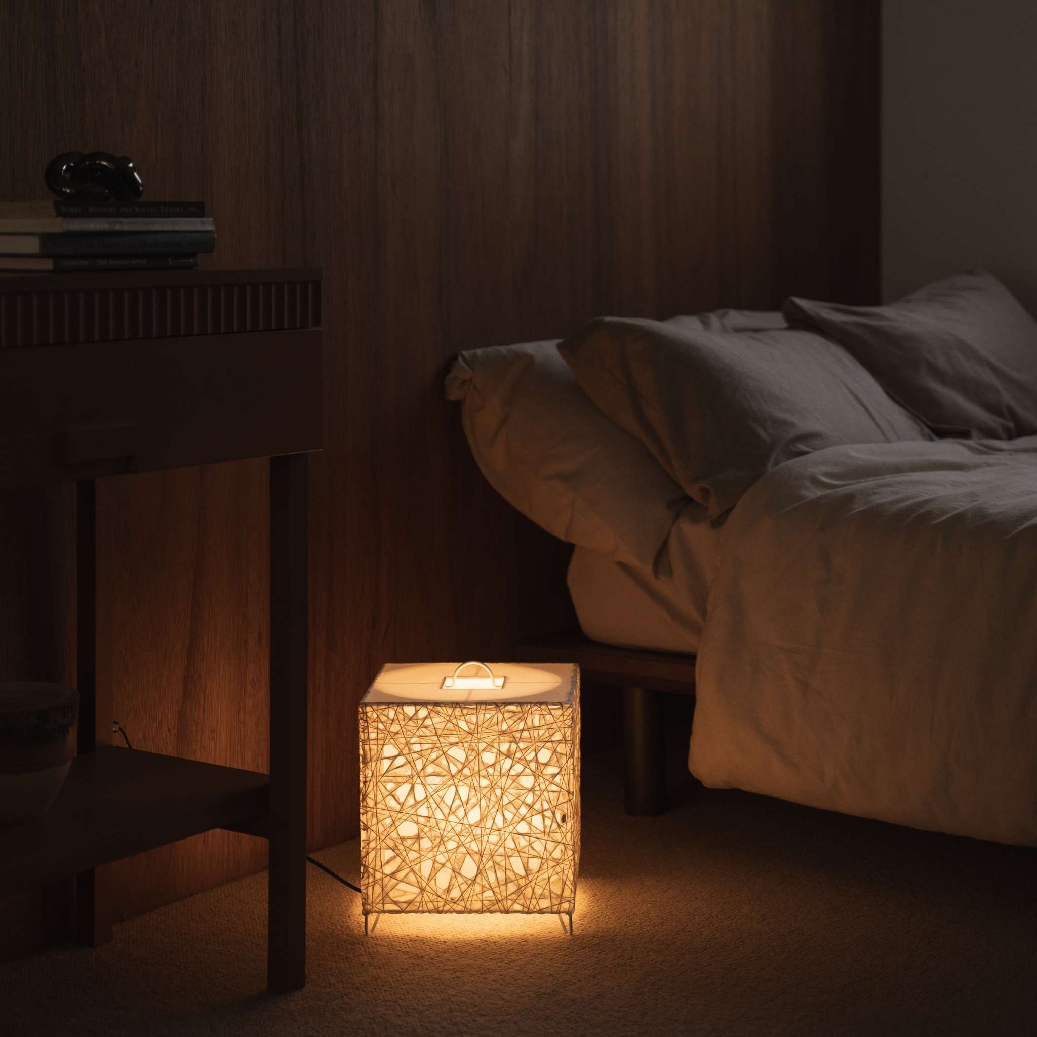 "Lampe japonaise en papier Me-Look 03 en forme de cube, blanc, diffusant une lumière chaude sur une table de chevet en bois dans une chambre japonaise."