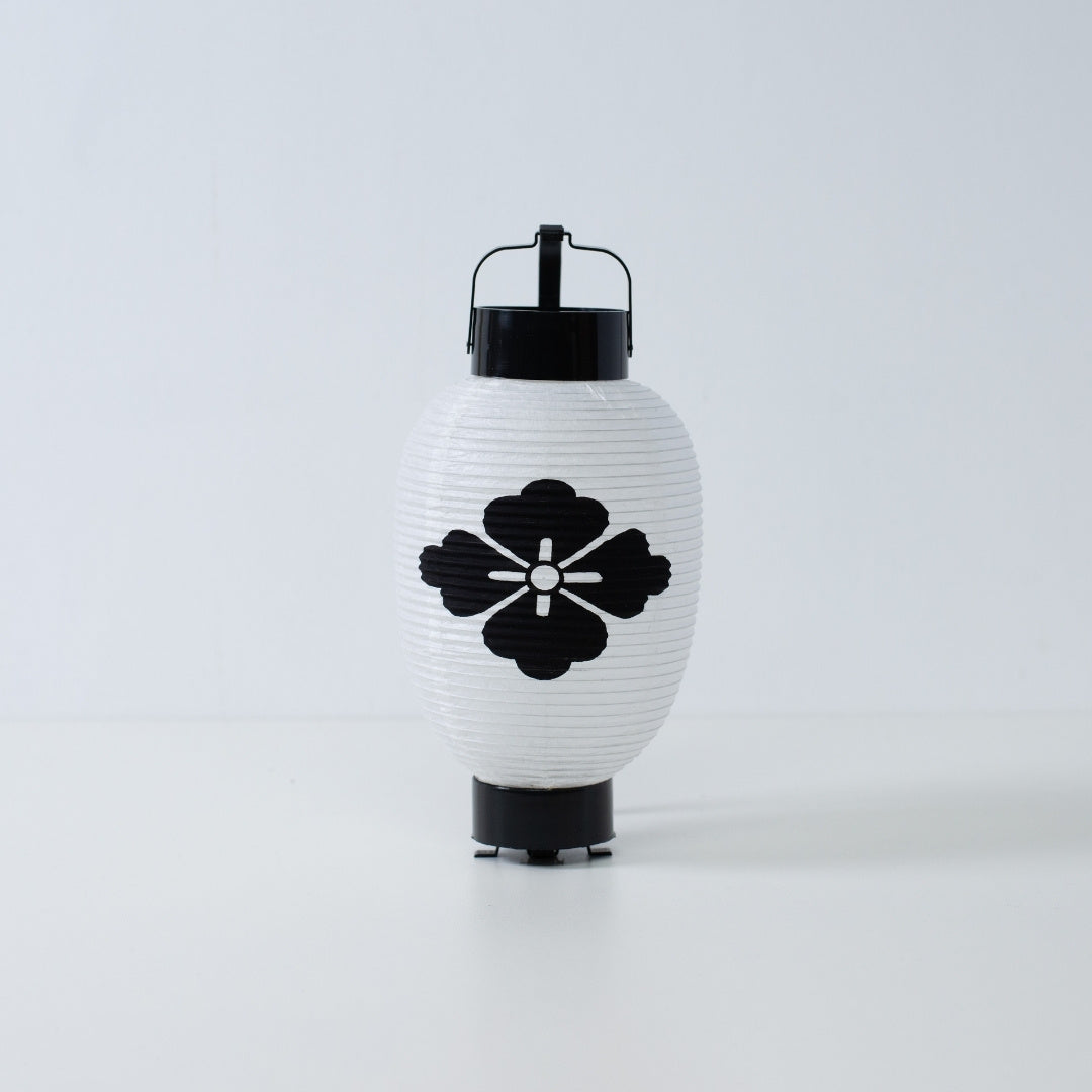 "Een witte Japanse papieren lantaarn met een handgeschilderd zwart bloemvormig hanabishi-embleem op een witte achtergrond."