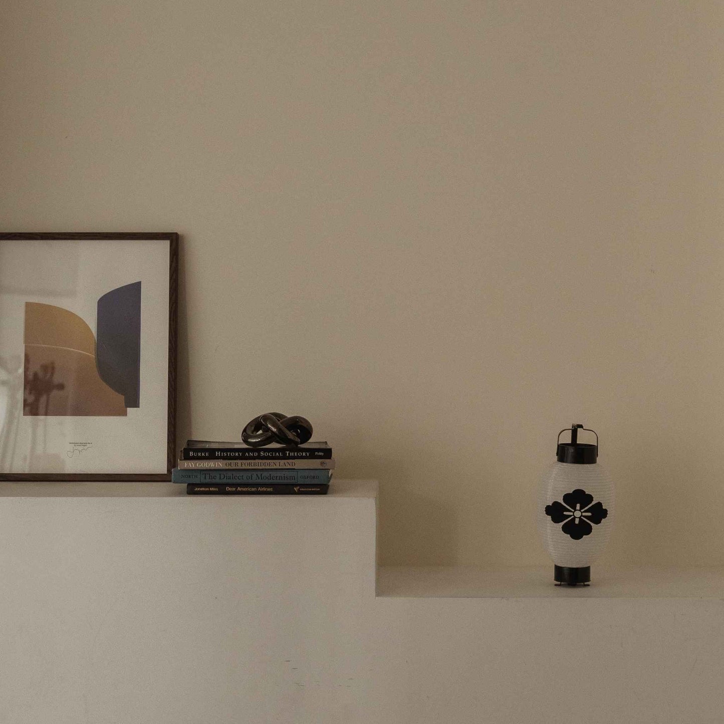 "Minimalistische witte plank met naast een ingelijst kunstwerk een Japanse papieren lantaarnbloem in diamantvorm."