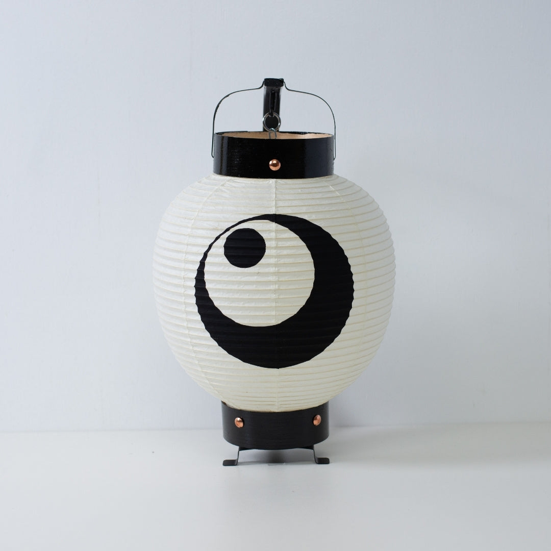 Een kleine Japanse papieren lantaarn met een handgeschilderd zwart maan- en sterrenwapen, staand op een wit oppervlak.