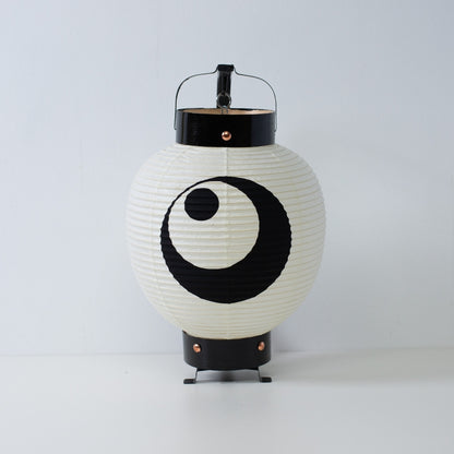 Een kleine Japanse papieren lantaarn met een handgeschilderd zwart maan- en sterrenwapen, staand op een wit oppervlak.