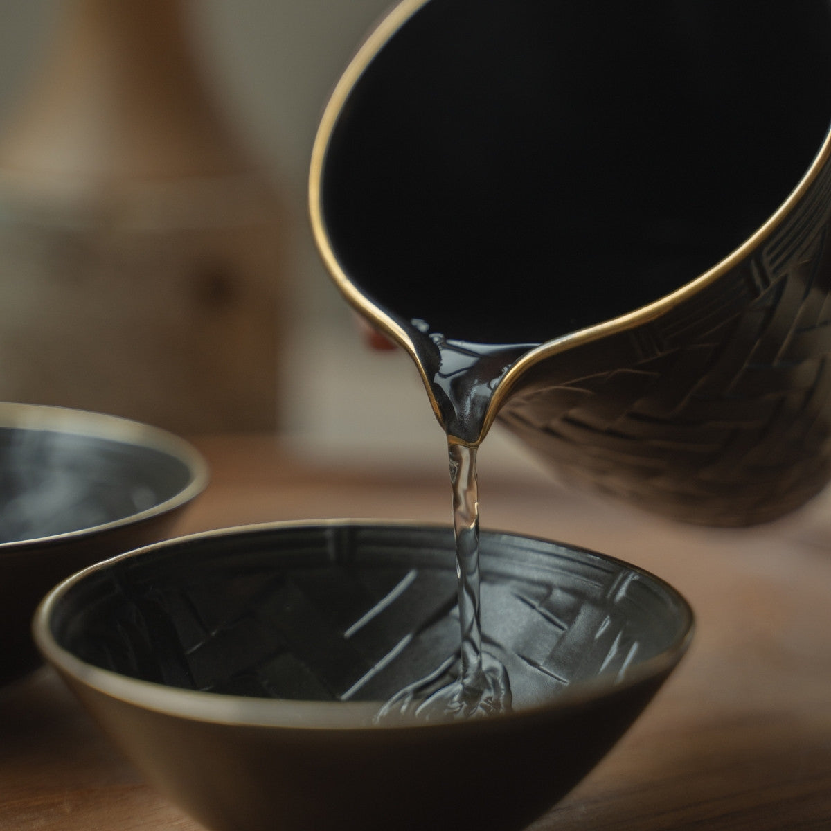 Hand pouring clear sake from a black Japanese sake pitcher into a dark guinomi cup, elevating a special dinner.