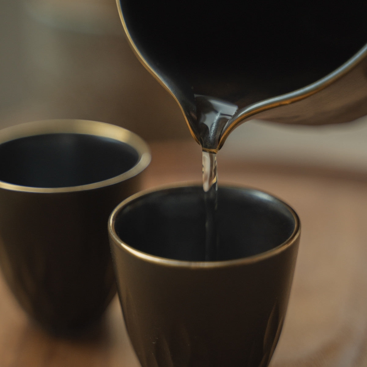 Gros plan d'un saké transparent versé d'une carafe à saké noire dans une tasse à saké à bordure dorée, rehaussant un moment de dîner luxueux.