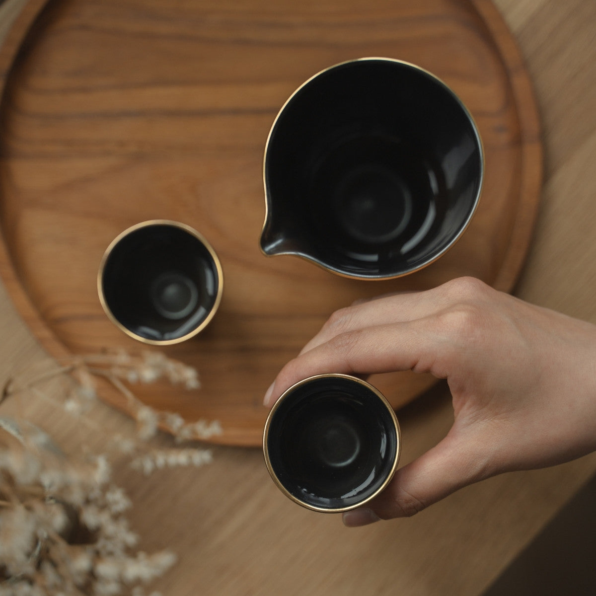 Main tenant une tasse à saké noire révélant son intérieur argenté sur un plateau en bois, se préparant pour un rituel de saké en soirée empreint de pleine conscience.