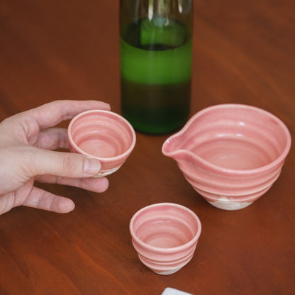 A person holds a pink Japanese Kyo-yaki sake cup (ochoko) above a wood table, with a sake bottle and katakuchi carafe nearby.