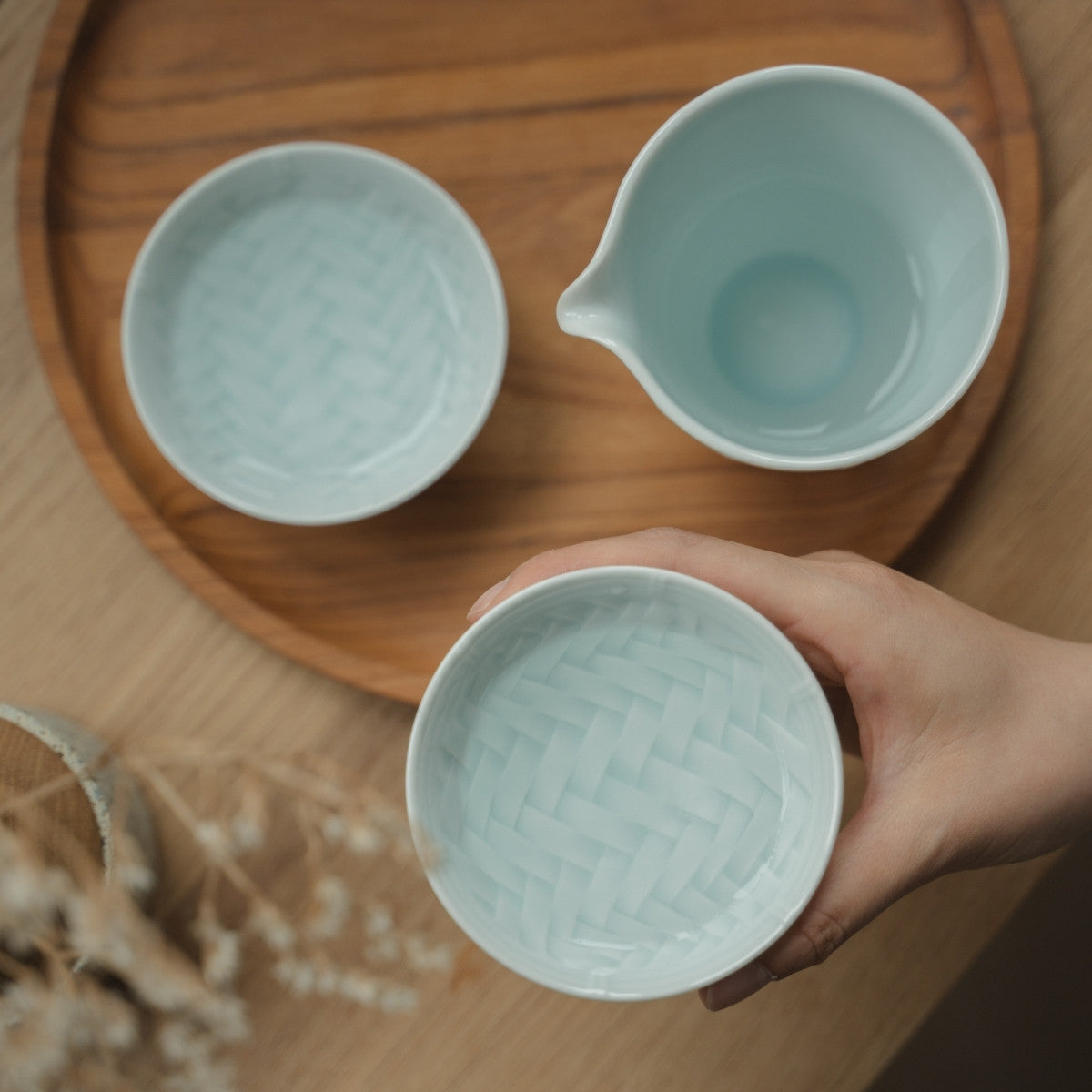 Main tenant une coupe à saké guinomi en porcelaine japonaise Arita bleu glacier au-dessus d'un plateau, invitant les amateurs de saké à profiter d'une soirée tranquille.