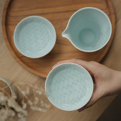 Main tenant une coupe à saké guinomi en porcelaine japonaise Arita bleu glacier au-dessus d'un plateau, invitant les amateurs de saké à profiter d'une soirée tranquille.
