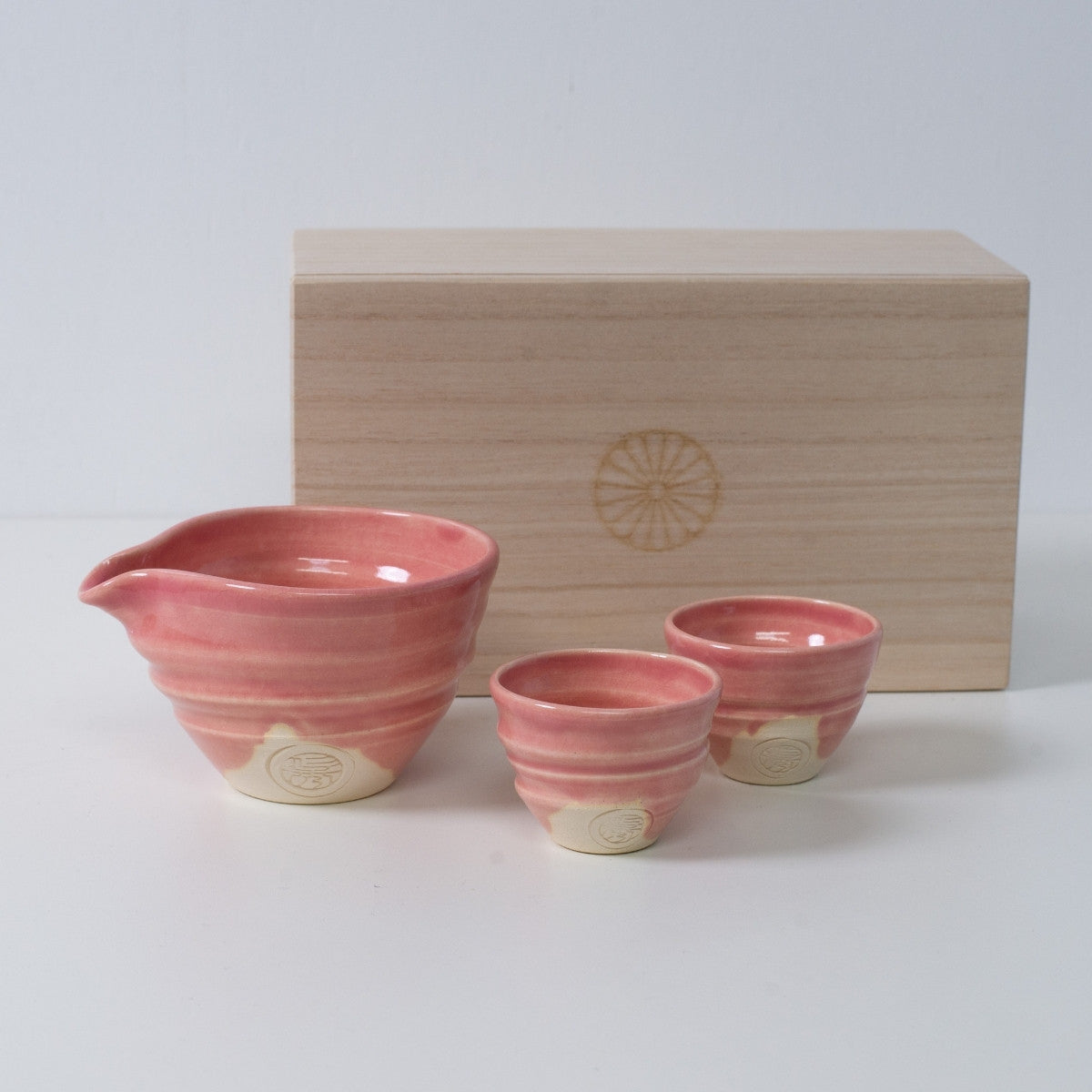Pink Japanese sake set "Omuro Cherry" pottery with a sake carafe and two cups, next to a wooden box with a chrysanthemum crest.
