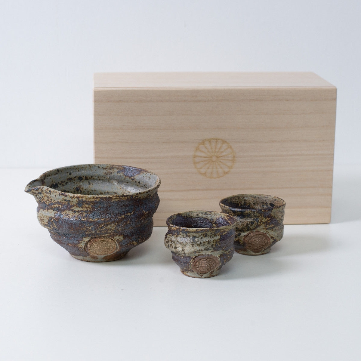 Rustik brun japansk sake-sæt "Sakigake" keramik med en sake-karaffel og to kopper, ved siden af en trækasse med et krysantemum-emblem.