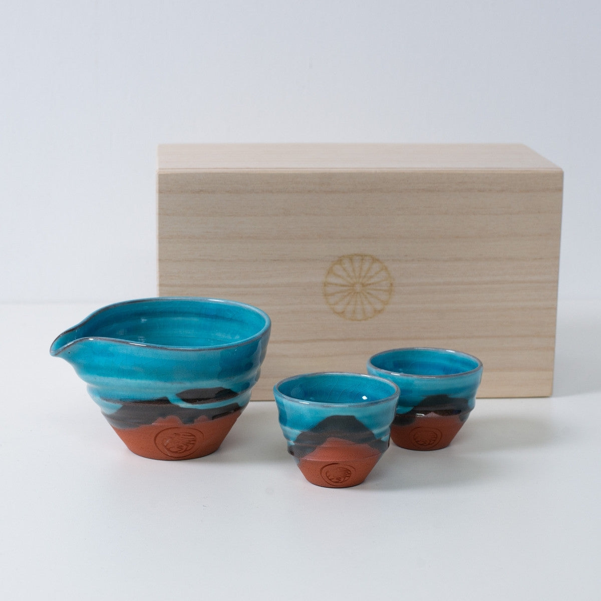 Turkis japansk sake-sæt "Soku Blue" keramik med en sake-karaffel og to kopper, ved siden af en trækasse med et krysantemum-emblem.