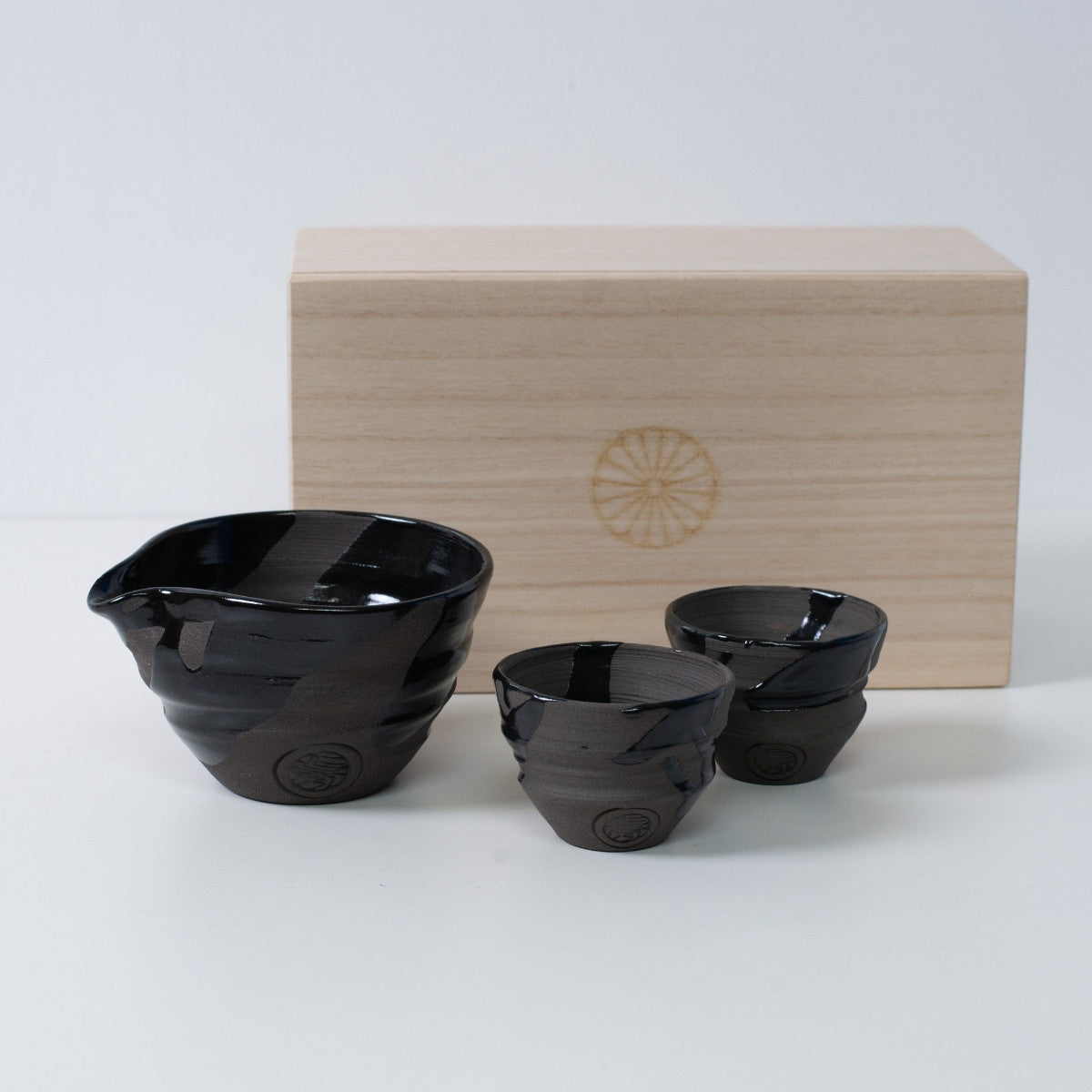 Japansk sake-sæt "Sumi Black" i keramik med en sake-karaffel og to kopper, ved siden af en trækasse med et krysantemum-emblem.