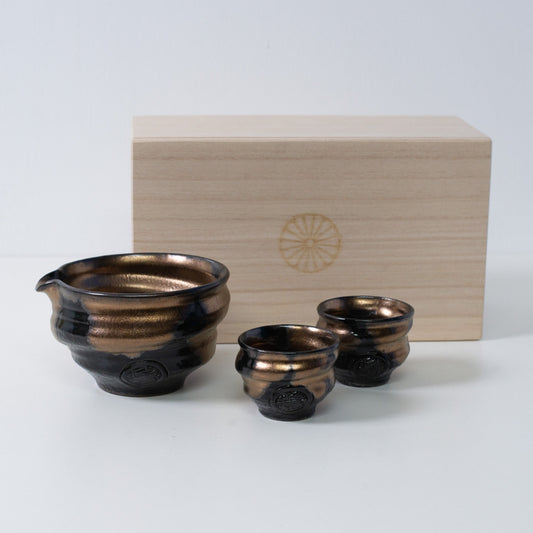 Gold and black Japanese sake set "Zen" pottery with a sake carafe and two cups, beside a wooden box with a chrysanthemum crest.