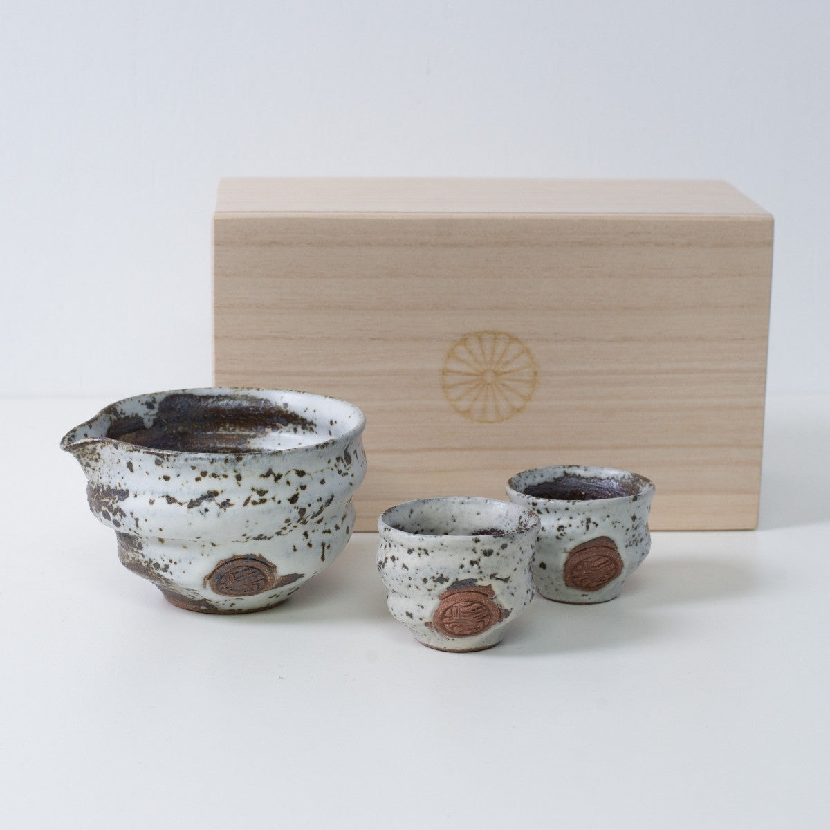 Brown and white Japanese sake set "Zui" pottery with a sake carafe and two cups, next to a wooden box with a chrysanthemum crest.