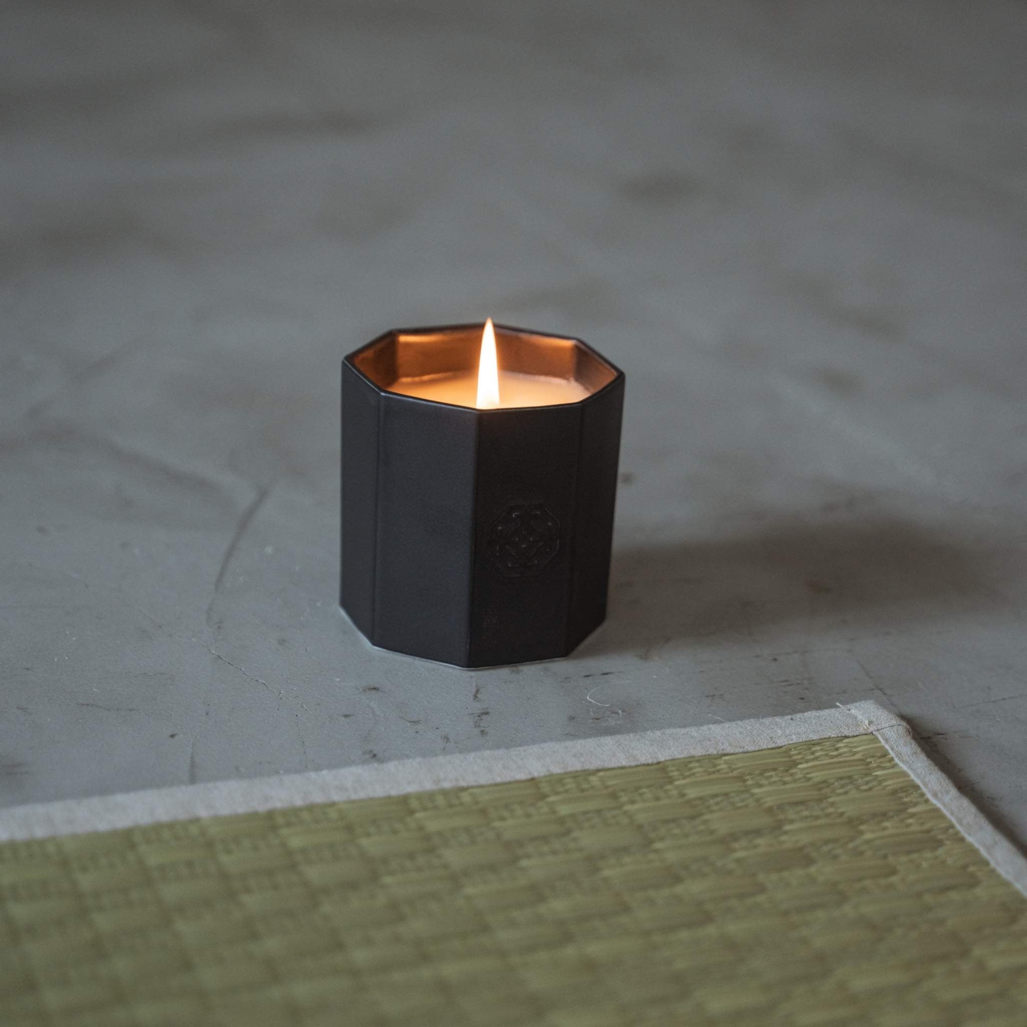 Sort arita-keramik duftlys, der brænder med en varm flamme på et gråt gulv ved siden af en traditionel tatami-måtte.