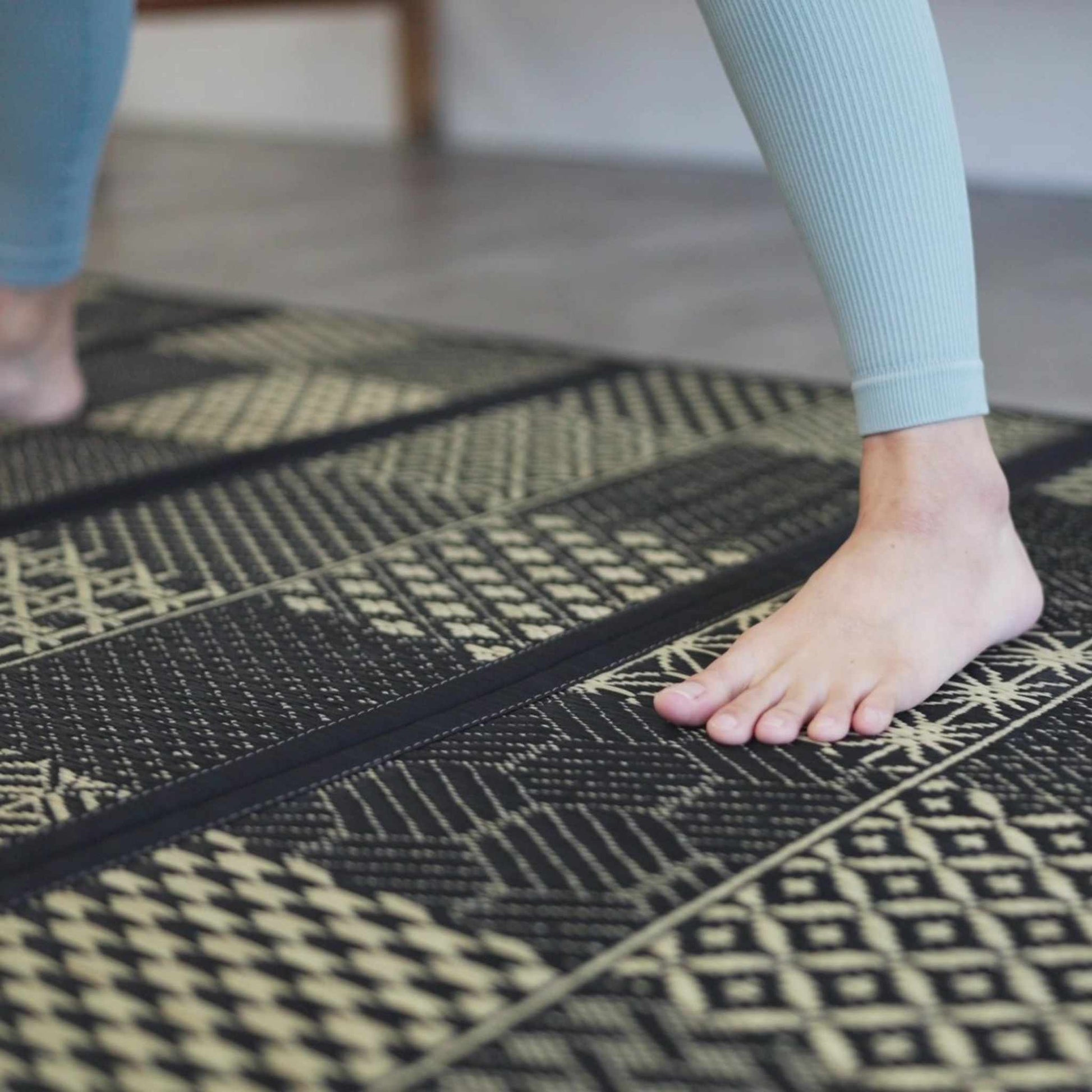 Close up of feet on a black patterned tatami mat showing the woven texture ideal for yoga and barefoot living.