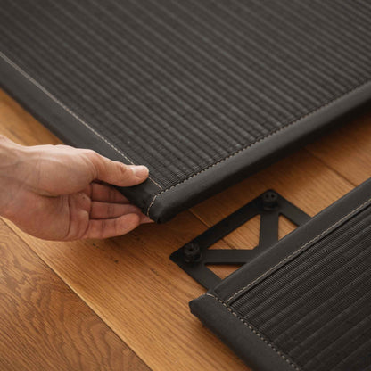 Hände beim Zusammensetzen von zwei schwarzen, quadratischen Tatami-Matten auf einem Holzboden, was den einfachen Aufbau für einen Zen-Bereich demonstriert.