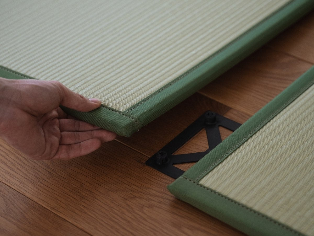 Hände verbinden zwei grüne Tatami-Mattenpaneele auf einem Boden, die den einfachen Aufbau für einen individuellen japanischen Raum demonstrieren.