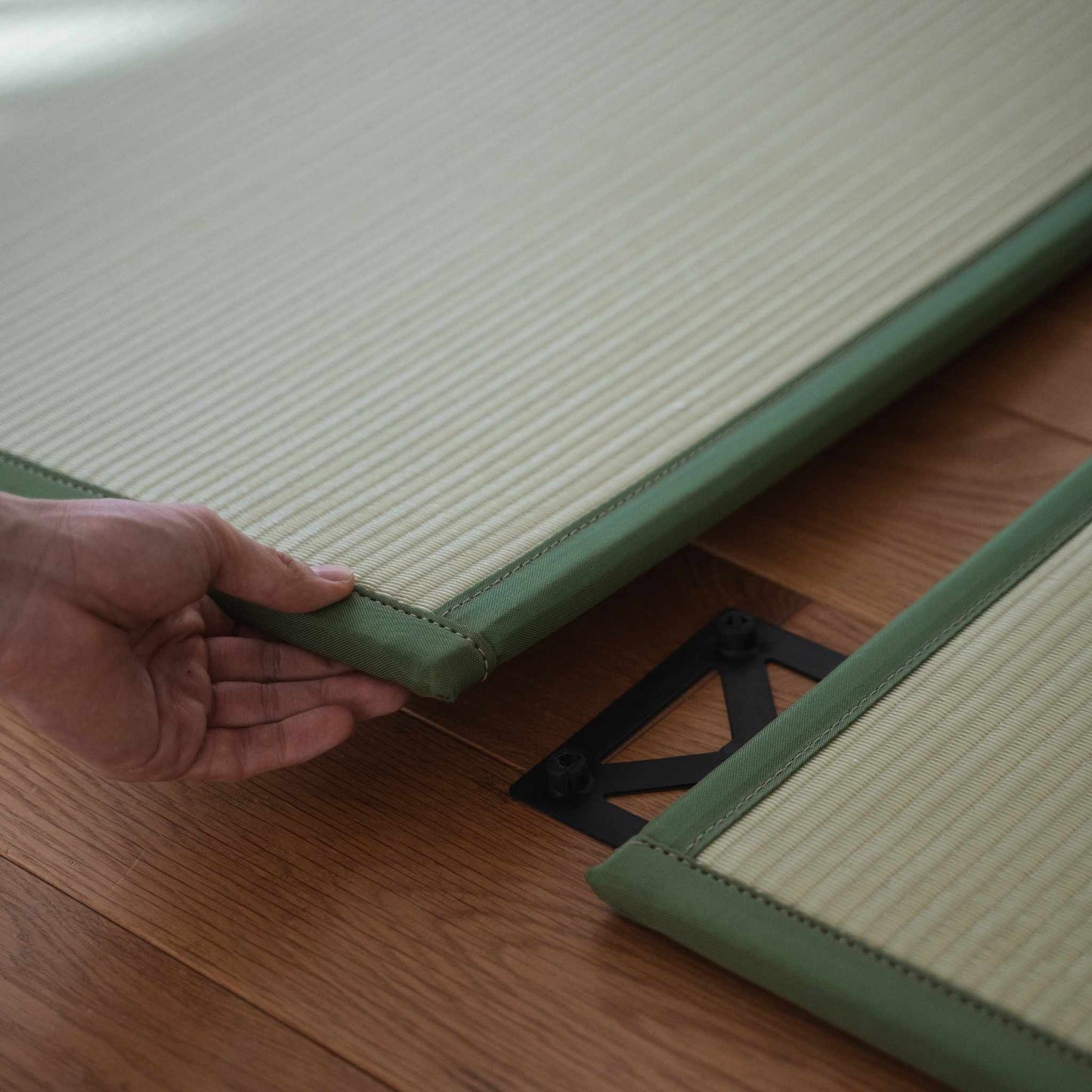 Handen die twee groene tatamimatpanelen op een vloer verbinden, wat de eenvoudige installatie van een aangepaste Japanse ruimte demonstreert.
