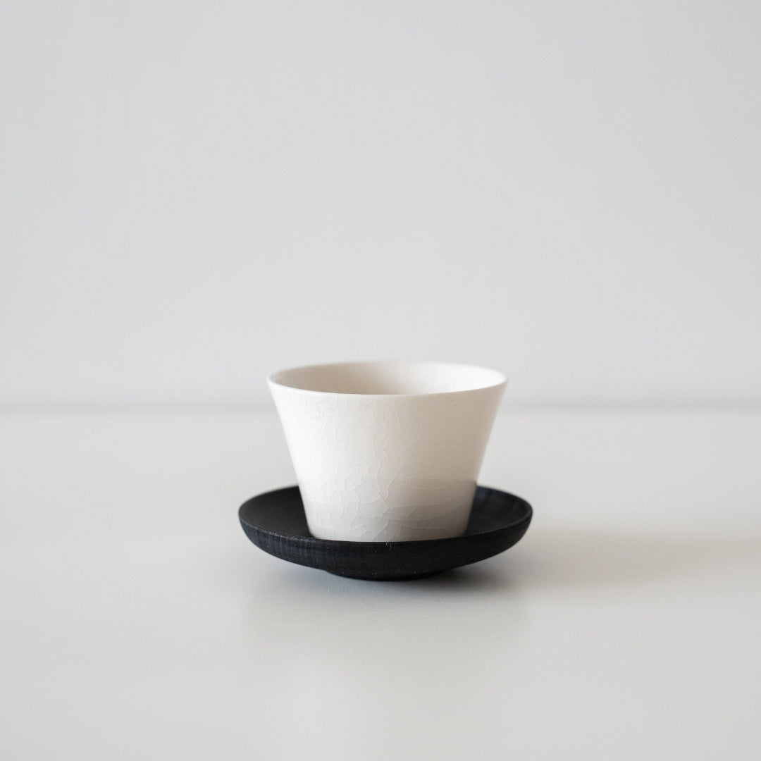 Eine einzelne, hochbeinige japanische Teetasse auf einer dunklen Untertasse, die die elegante Porzellanoberfläche im Wabi-Sabi-Stil hervorhebt.