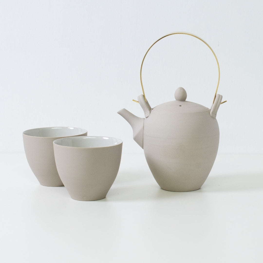 Moderne japansk tesæt med en hvid ler kyusu tekande med messinghåndtag ved siden af to buede tekopper i f-form