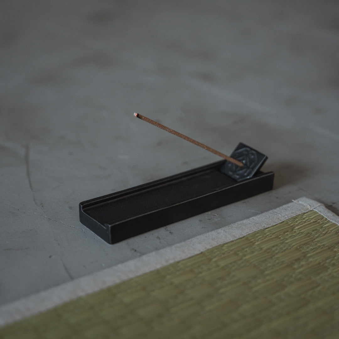 Ein japanisches Hinoki-Räucherstäbchen brennt in einem schwarzen Porzellanhalter auf einer Zen-Tatami-Yogamatte für Wellness.