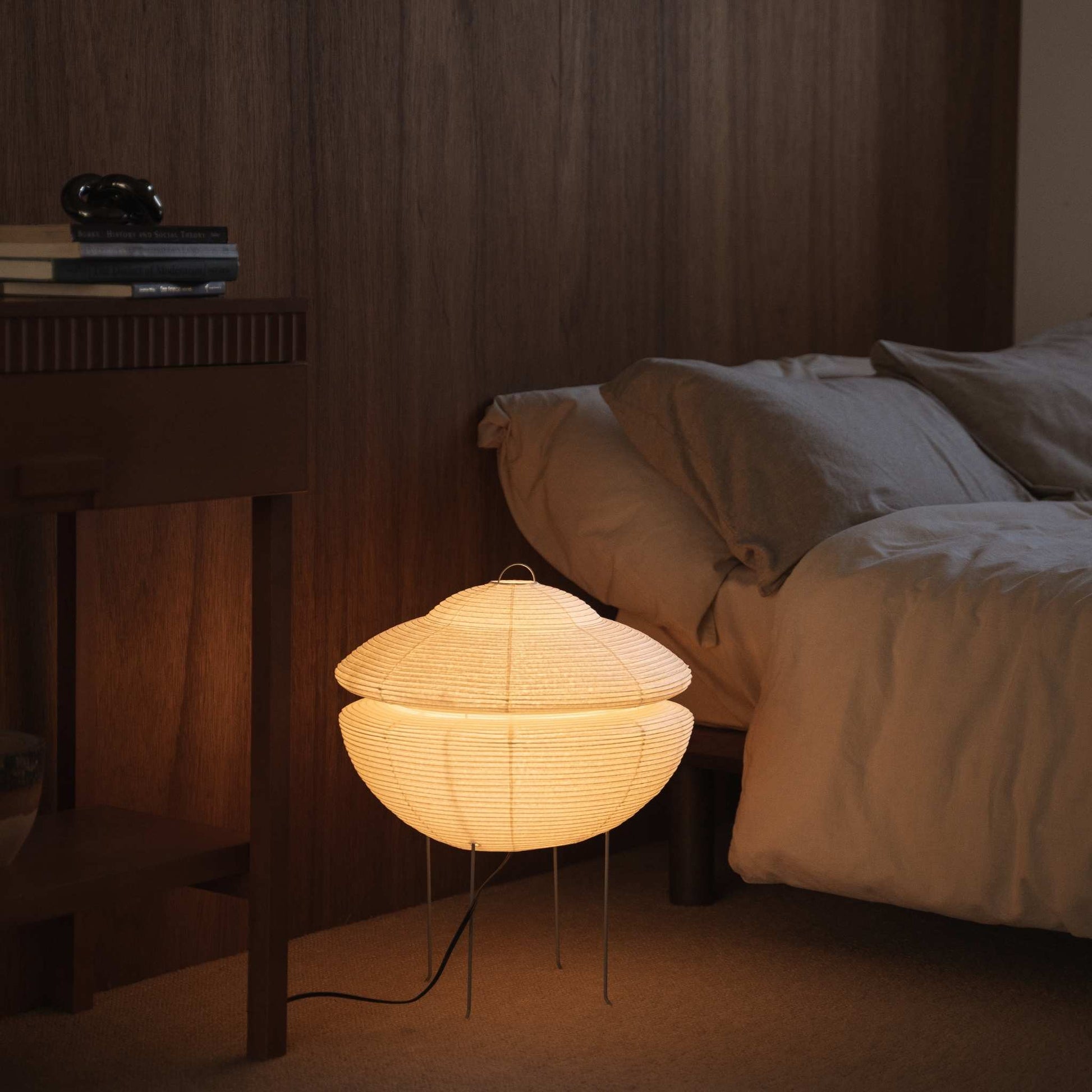 "Lampe sur pied japonaise illuminée en papier Gemini 01 diffusant une lumière douce et chaude, posée au sol à côté d'un lit dans une chambre japonaise"