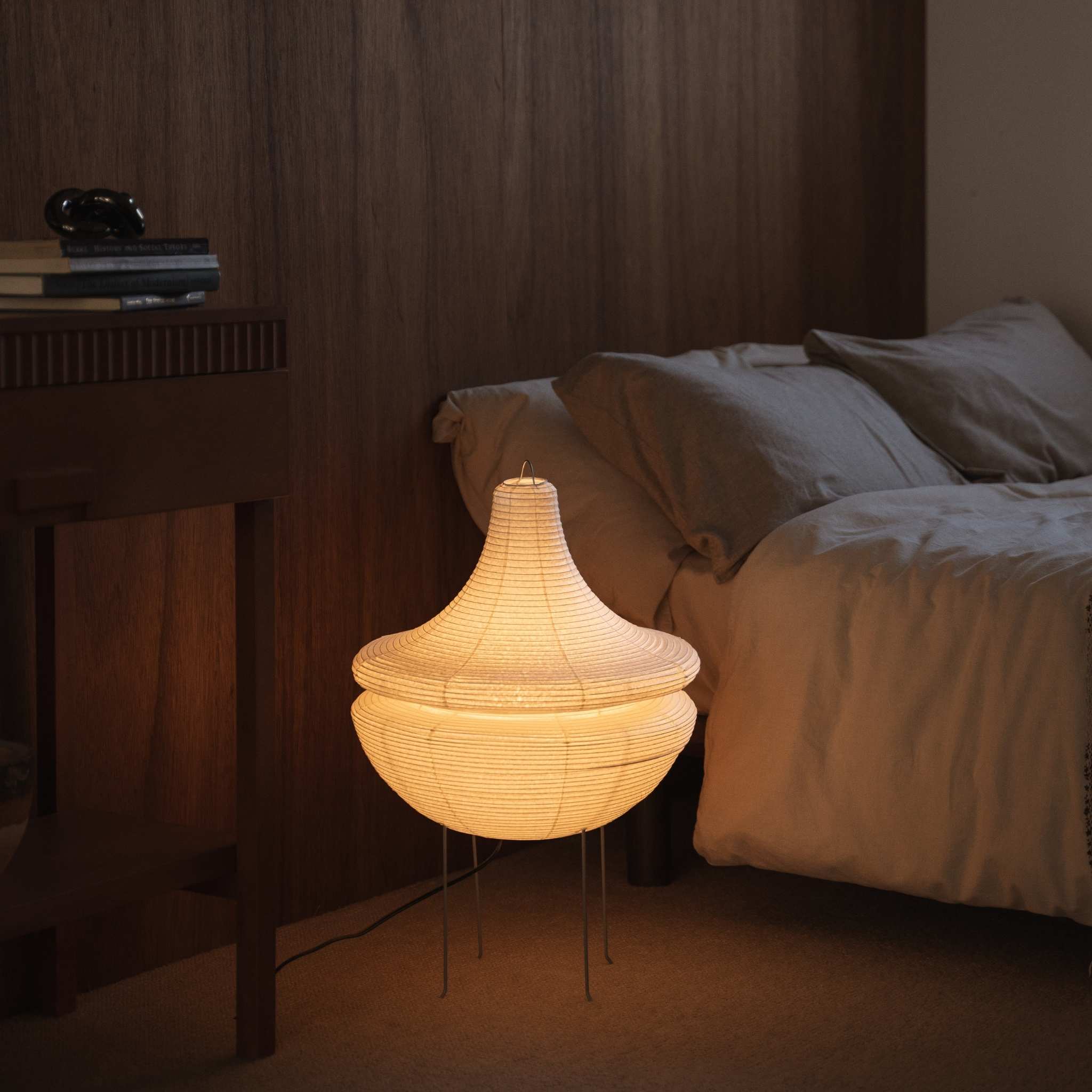 "Lampe sur pied japonaise illuminée Gemini 02 en papier, diffusant une lumière douce et chaude, posée au sol à côté d'un lit dans une chambre japonaise"